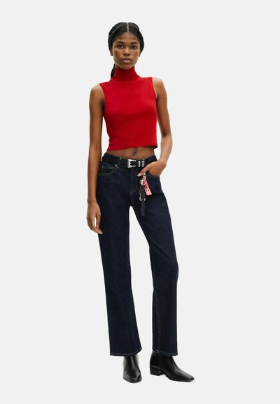 Top sin mangas de color rojo con cuello alto, combinado con jeans rectos de pierna oscura. Cinturón negro con acentos de llavero. Botines negros.