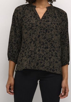 Vrouw draagt donkergroen oliën blouse met zwart bloemenpatroon en driekwartmouwen, gecombineerd met zwarte broek tegen een effen achtergrond.