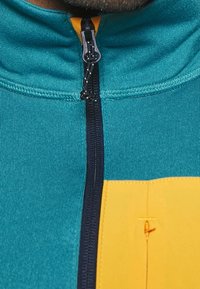 Giacca a zip color teal con dettagli arancioni sulla cerniera. Presenta un tessuto testurizzato e una grande toppa gialla. La cerniera scorre verticalmente lungo il davanti.