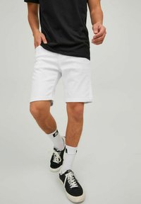Pantalones cortos de mezclilla blancos con dobladillos enrollados, combinados con una camiseta negra y zapatillas negras. Los calcetines son blancos con un logo. Enfoque en un estilo casual.