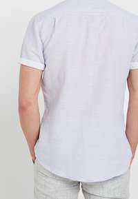 Camicetta azzurro chiaro a maniche corte con tessuto testurizzato. Presenta un orlo curvo e polsini ripiegati. La parte posteriore include due cuciture a yoke.