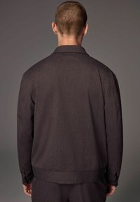 Dark brown jacket with a textured fabric, button cuffs, collar, and a cropped hem. Displayed from the back against a gray background.