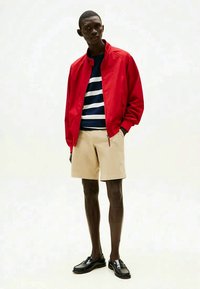 Male model wearing a red jacket, navy and white striped shirt, beige shorts, and black loafers, standing with one hand in pocket.