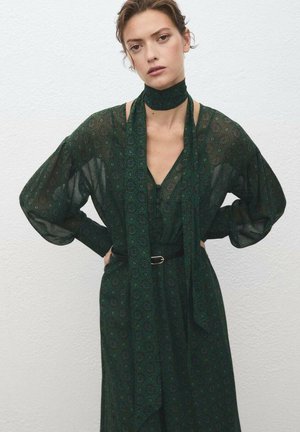 Abito verde a fantasia con scollatura profonda a V, maniche lunghe e colletto legato. Presenta una cintura in vita e una texture leggera e fluttuante.