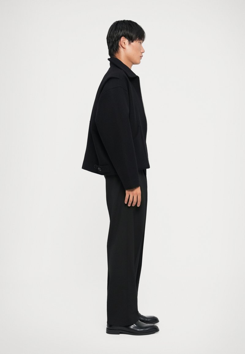 Black cropped wool jacket with a high collar and side button detail, paired with black trousers and leather shoes, displayed from the side.