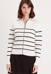 Cardigan rayé en maille côtelée, présentant des rayures horizontales noires sur un fond blanc, fermeture éclair et col. Manches et bas texturés.