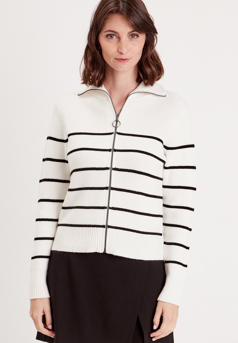 Cardigan rayé en maille côtelée, présentant des rayures horizontales noires sur un fond blanc, fermeture éclair et col. Manches et bas texturés.