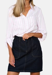 Kvinna klädd i en vit skjorta med rosa ränder och uppvikta ärmar, instoppad i en mörkblå denimkjol med framfickor.