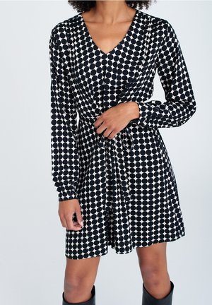 Robe à pois noir et blanc avec manches longues, décolleté en V, taille ajustée et un détail à nouer à la taille. Texture de tissu lisse.