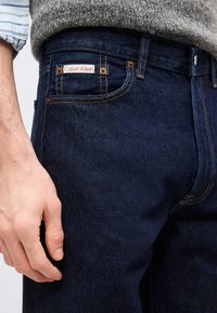 Κοντινό πλάνο ενός ατόμου που φορά σκούρα μπλε τζιν Calvin Klein και γκρι πουλόβερ, με το αριστερό χέρι χαλαρό στο πλευρό του.