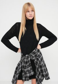 Juodas megztinis su aukštu apykakliu ir deimantų raštais, derinamas su juoda ir pilka languota marška, surišta aplink juosmenį.