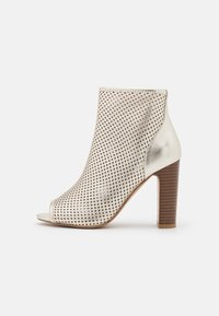 Sandały typu ankle boot z odkrytymi palcami w metaliczno-beżowym kolorze z perforowanym wzorem. Posiadają masywny, drewniany obcas oraz gładkie akcenty teksturowe.