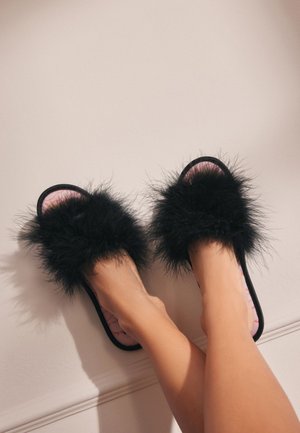 Fekete szőrös papucsok plüss anyagból, nyitott orrú kialakítással és puha rózsaszín bélésbelsővel. A talp fekete, minimális díszítéssel.