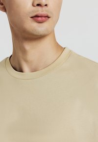 Primo piano della parte inferiore del viso e del collo di una persona che indossa una maglietta beige a girocollo, rivolta leggermente di lato su uno sfondo neutro.