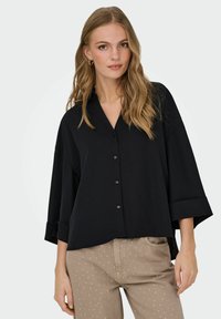 Sort oversized skjorte med en klippet krave, frontknapper og brede ærmer. Båret med lysebrune bukser med hvide prikker.