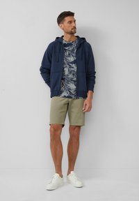 Granatowa rozpinana bluza z kapturem nałożona na niebieską i białą koszulę w tropikalny wzór, w połączeniu z beżowymi szortami i białymi sneakerami. Gładkie, casualowe tekstury materiałów.