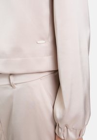 Beige Satin-Outfit mit lockerer Passform. Merkmale sind geraffte Ärmel, ein dezentes Logotag nahe dem Saum und eine samtige Textur.
