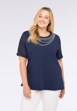 Donna sorridente con lunghi capelli biondi che indossa una maglietta blu navy con dettagli in perle e maniche trasparenti, abbinata a pantaloni bianchi, su sfondo bianco.