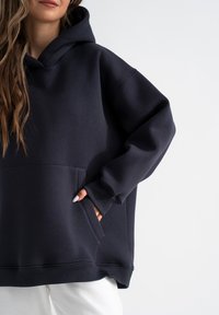 Marineblaue oversized Kapuzenjacke aus weichem Material, mit einer Kängurutasche und gerippten Bündchen. Schlichtes, minimalistisches Design.