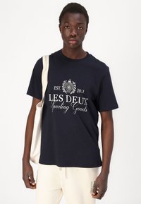 Tums navy zila krāsā ar baltu grafiku, uz kuras ir teksts "LES DEUX Sporting Goods" un apļveida emblēma. Valkāta ar bēšu auduma somu.