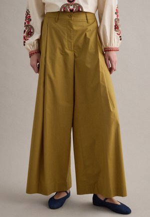 Pantalon large taille haute couleur moutarde associé à un chemisier crème avec des motifs brodés rouges et marron sur les manches et la poitrine.