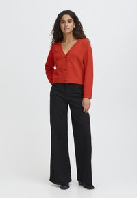 Cardigan rosso lavorato a maglia con scollatura a V profonda e chiusura a toggle frontale, abbinato a pantaloni neri a vita alta e ampi e stivali neri alla caviglia.