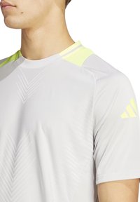 Lichtgrijs sportshirt met korte mouwen en gele accenten. Heeft gestructureerde patronen en een ronde halslijn. Gemaakt van ademende stof.