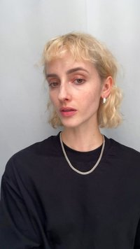 Collier en maille argentée reposant sur une chemise noire, avec une paire de boucles d'oreilles rondes et brillantes en argent visibles. Des cheveux blonds courts et ondulés encadrent le visage.