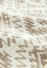 Textil, amely világosbarna geometriai mintával rendelkezik egy krémszínű háttéren, textúrált felülettel és bordázott megjelenéssel.