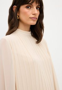 Blusa lunga plissettata in un morbido beige con un collo alto a coste. Texture trasparente con sottili linee verticali, abbinata a orecchini a cerchio dorati.