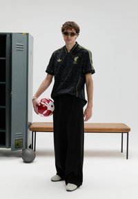 Fekete mintás focimez sárga díszítéssel, laza fekete nadrág és fehér cipők. Egy piros-fehér focilabdát tart. Minimalista háttér.