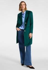 Grüner langer Blazer mit goldenen Knöpfen über einem hellblau gestreiften Hemd, kombiniert mit weit geschnittenen blauen Jeans und braunen Absätzen. Glatte Textur.