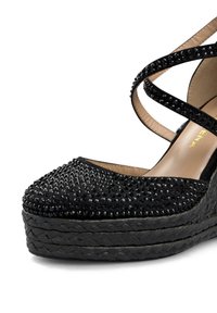 Sandale compensée noire avec une tige en tissu texturé ornée de strass, dotée de brides de cheville croisées et d'une plateforme tressée.