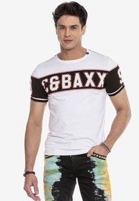 Witte en zwarte T-shirt met korte mouwen en opvallende rode en witte letters. Model draagt tie-dye jeans met decoratieve studs langs de tailleband.