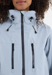 Helleblaue, wasserdichte Jacke mit hohem Kragen, schwarzen Reißverschlüssen und verstellbaren Zugbändern. Verfügt über zwei Reißverschluss-Fronttaschen.