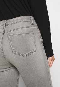 Persona wearing fitted light gray denim jeans and a black long-sleeve top, mostrando el bolsillo trasero y la mano derecha descansando sobre el muslo.