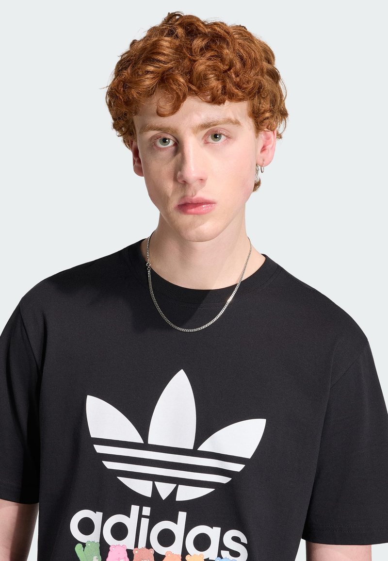 Junge Person mit lockigen roten Haaren, trägt ein schwarzes Adidas-T-Shirt mit weißem Logo und eine silberne Halskette, blickt nach vorne vor einfachem Hintergrund.