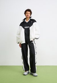 Čierno-biely Adidas teplákový komplet s bundou na zips, obsahujúcou veľkú kapucňu a športový dizajn. Textúrované čierne nohavice majú bočné volány.