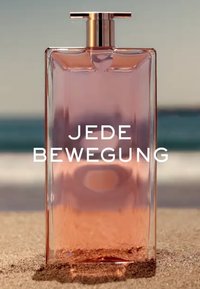 Doorzichtige glazen parfumfles met een rechthoekige vorm en een gepolijste gouden dop. De vloeistof verloopt van doorzichtig naar zachtroze en staat op zand.