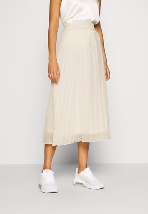 Vrouw draagt een wit mouwloos topje, een beige geplooide midirok en witte sportieve sneakers, staand tegen een effen achtergrond.