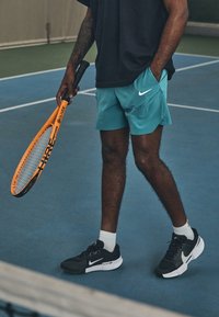Jugador de tenis con camiseta negra y pantalones cortos turquesa sostiene una raqueta naranja y negra; lleva zapatillas Nike en blanco y negro y calcetines blancos.