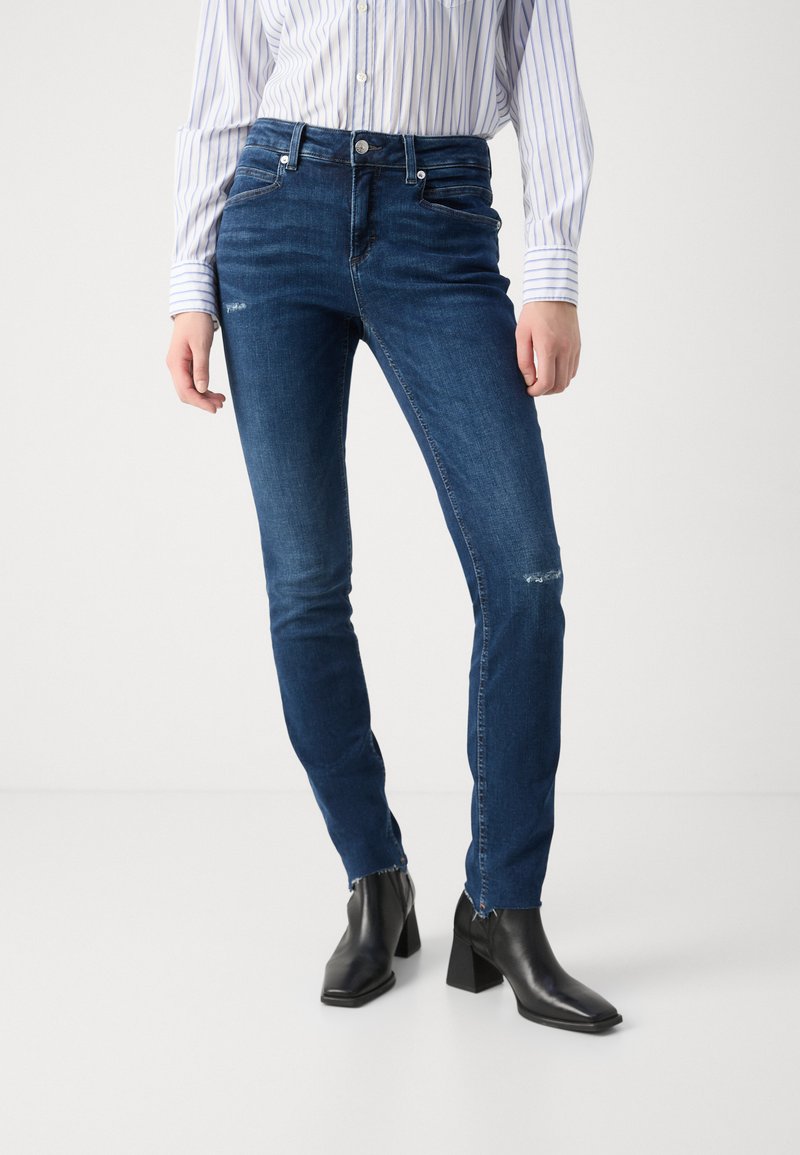 Jeans ajustados de color azul en denim con un ligero desgaste y desgarros en las rodillas, combinados con botas negras de tobillo y una camisa de botones a rayas.