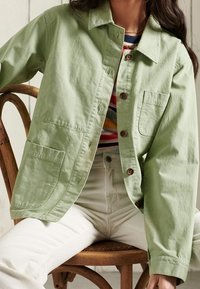 Person wearing light green buttoned jacket over striped shirt, sitting on wooden chair, with white pants.
