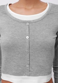 Cardigan gris court avec un col rond, doté d'une fermeture à boutons à l'avant et d'un t-shirt blanc visible en dessous de l'ourlet.