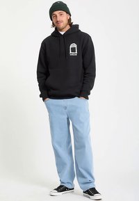 Sort hættetrøje med hvidt grafisk logo, kombineret med lyseblå afslappet-fit jeans og sorte sneakers, vist på en model.