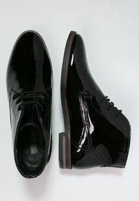 Zapatos de charol negro con un acabado brillante, que cuentan con un diseño de cordones, punta redondeada y un tacón grueso marrón.