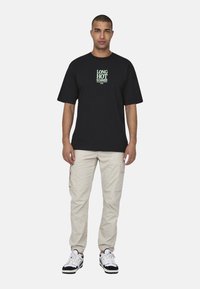 Czarny bawełniany t-shirt z zielonym napisem "DŁUGIE GORĄCE LATO" na klatce piersiowej, połączony z beżowymi spodniami cargo i białymi trampkami.