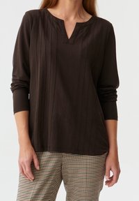 Haut marron à manches longues avec un col en V et une texture côtelée verticale. Porté avec un pantalon à carreaux à motifs. Finition en tissu lisse.