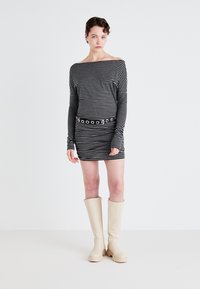 Schwarzes und weißes gestreiftes Kleid mit schulterfreier Ausschnittform und langen Ärmeln, verziert mit einem breiten Gürtel mit runden Ausschnitten. Beigefarbene Knief.-Stiefel.