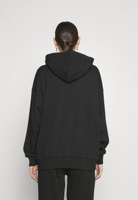 Musta ylikokoinen huppari, jossa suuri takahuppu, lasketut olkapäät, resorireuna ja teksturoitu kangas. Ei näkyviä logoja tai kuvioita.